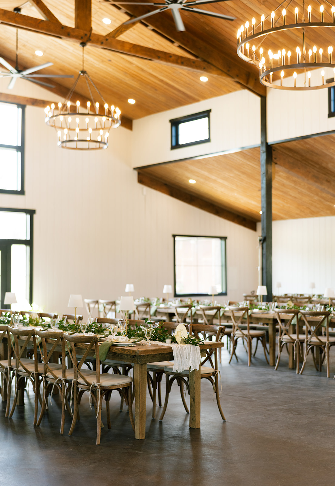 Long farm tables and chandeliers set for dinner inside the white barn